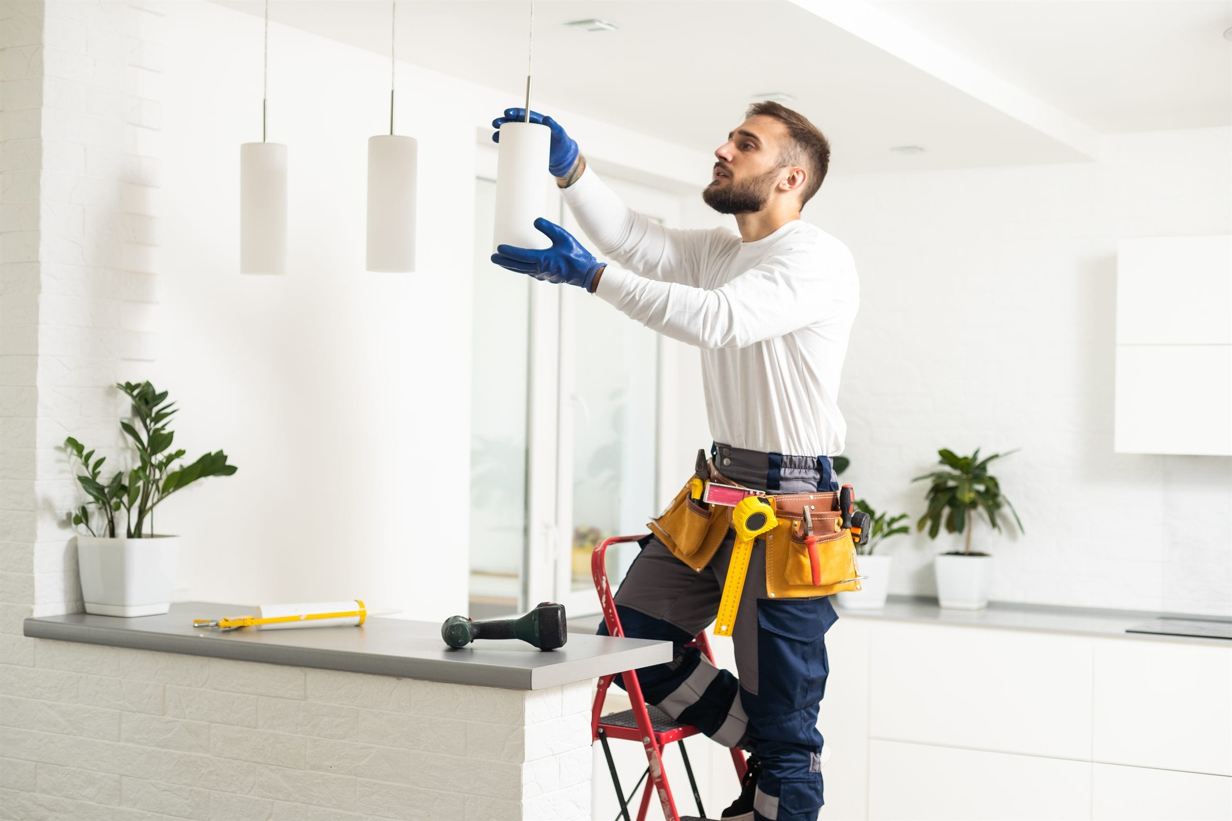 An electrician on a step ladder fitting a pendant light in a kitchen, tool belt on.