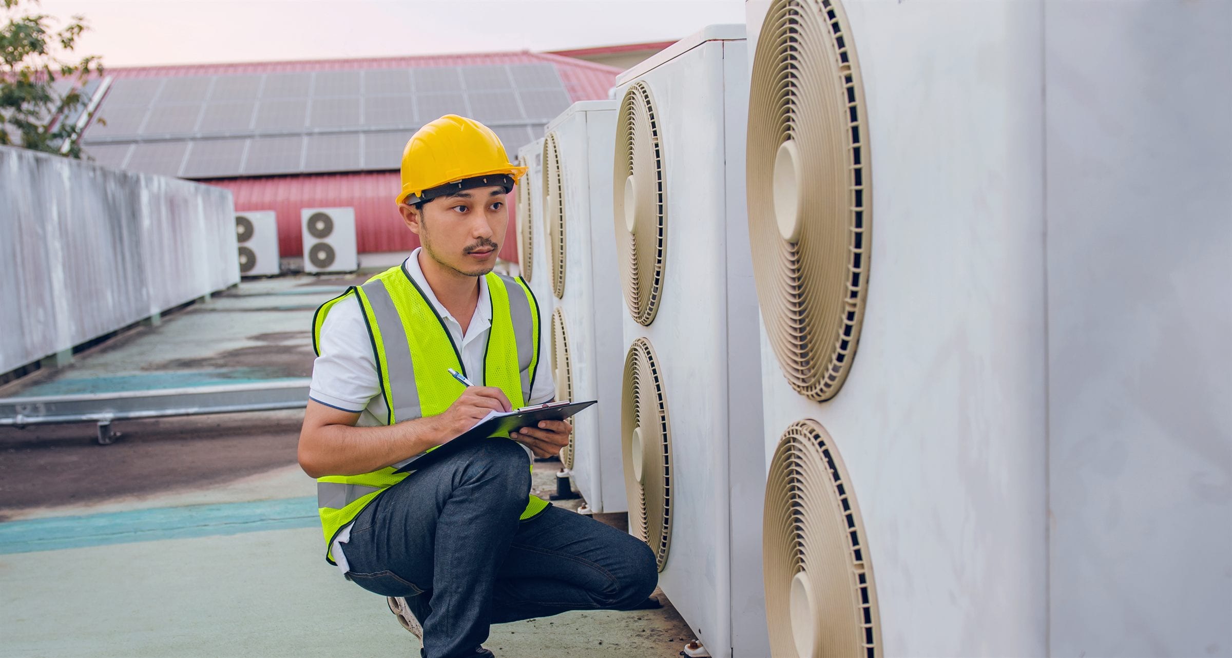 An HVAC technician logging a service check on a clipboard beside a bank of rooftop condenser units.