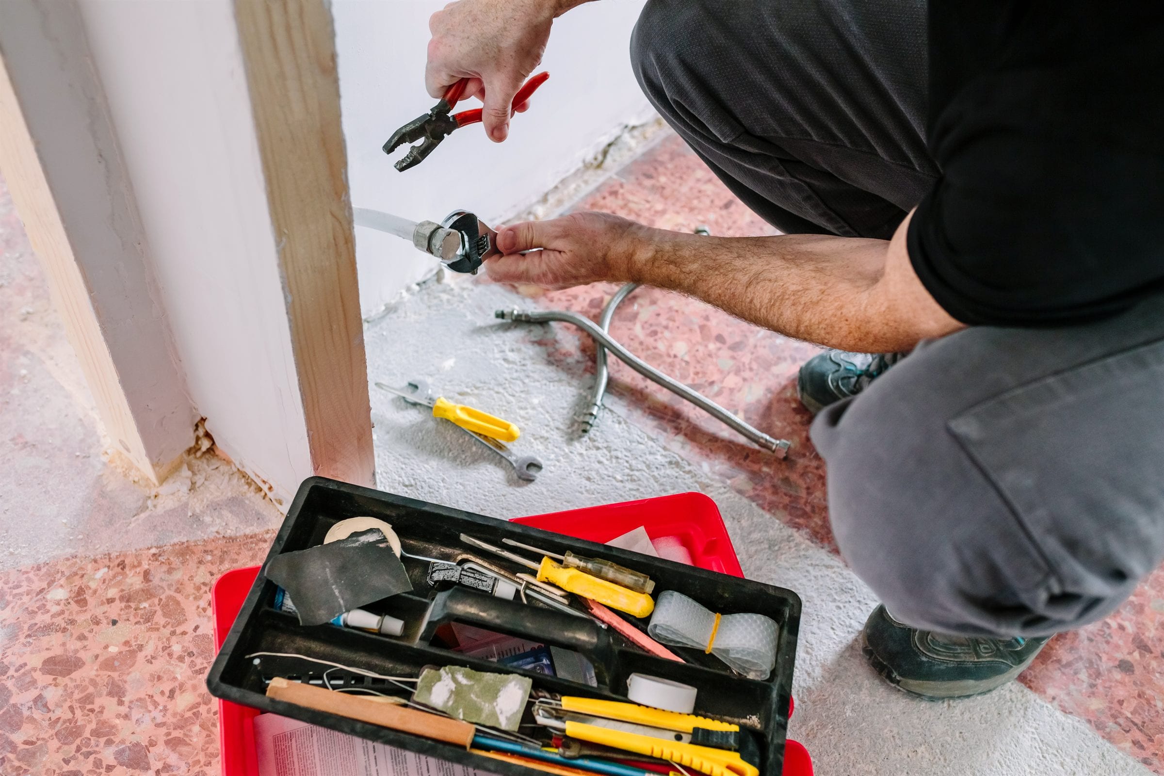 A plumber tightening a flexible supply line with an adjustable wrench, open toolbox on the floor beside him.