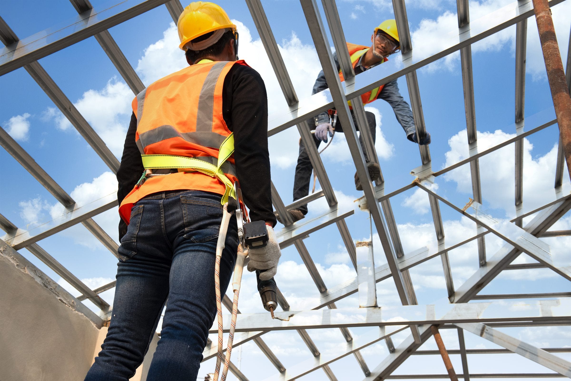 Two roofers in harnesses and high-vis fixing steel roof trusses against a blue sky.